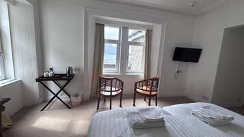 A hotel room with a bed, two chairs, a desk with a kettle, a TV, and a window with curtains.