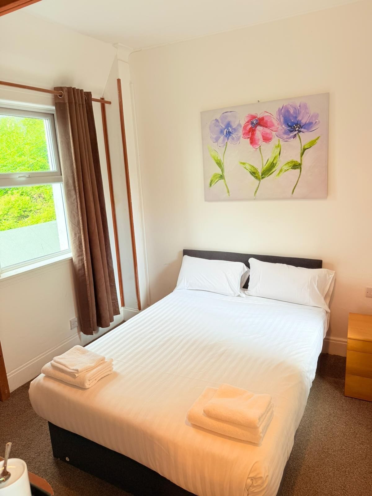 A neatly made bed with white linens and folded towels, a window with curtains, and a painting of flowers on the wall.