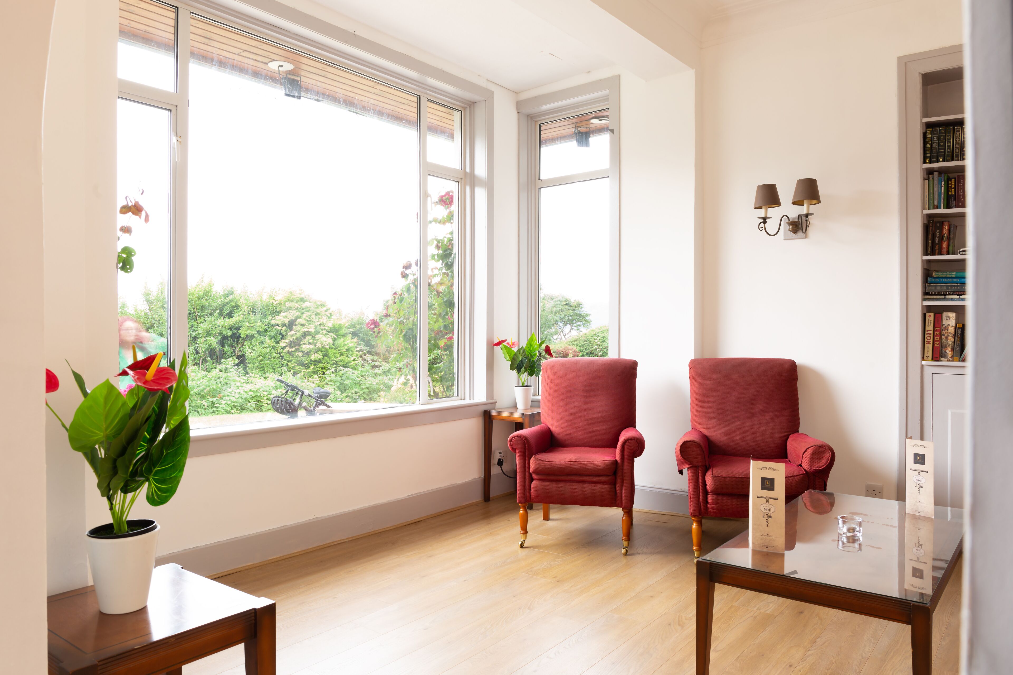 A room with a wooden floor, two red armchairs, a white table with a glass top, a potted plant, and a bookshelf.