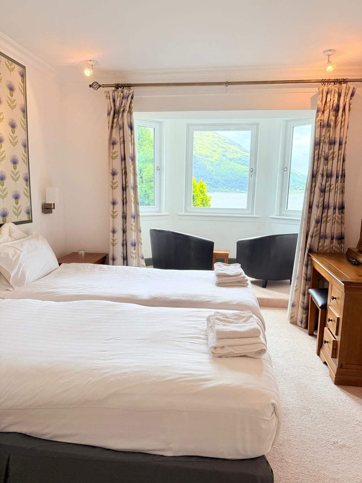 A hotel room with two beds, a desk, and a chair. There are windows with curtains, a view of greenery outside, and a lamp on the wall.