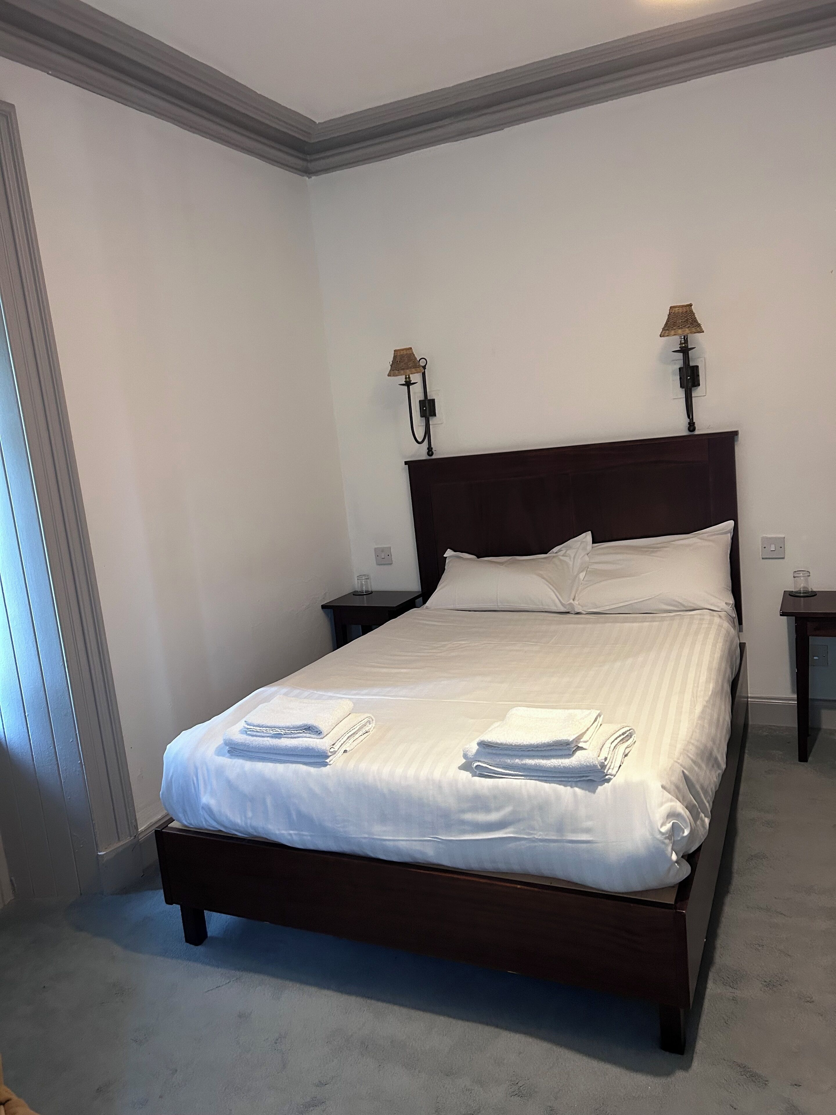 A neatly made bed with white linens and towels, two bedside lamps, and a dark wooden headboard in a room with white walls and a grey carpet.