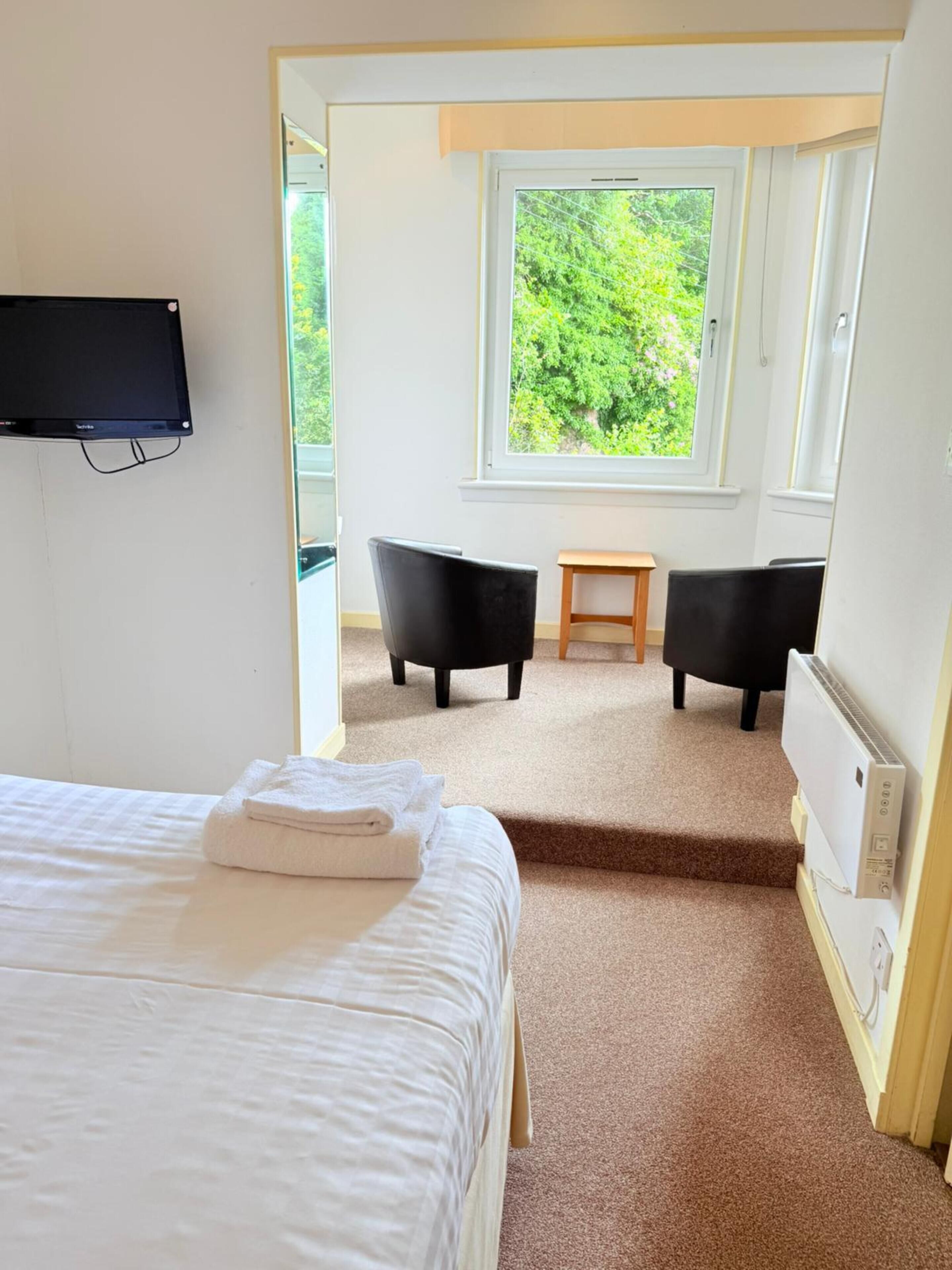A hotel room with a bed, a flat-screen TV mounted on the wall, a window with a view of greenery, two black chairs, a small wooden table, and a radiator.