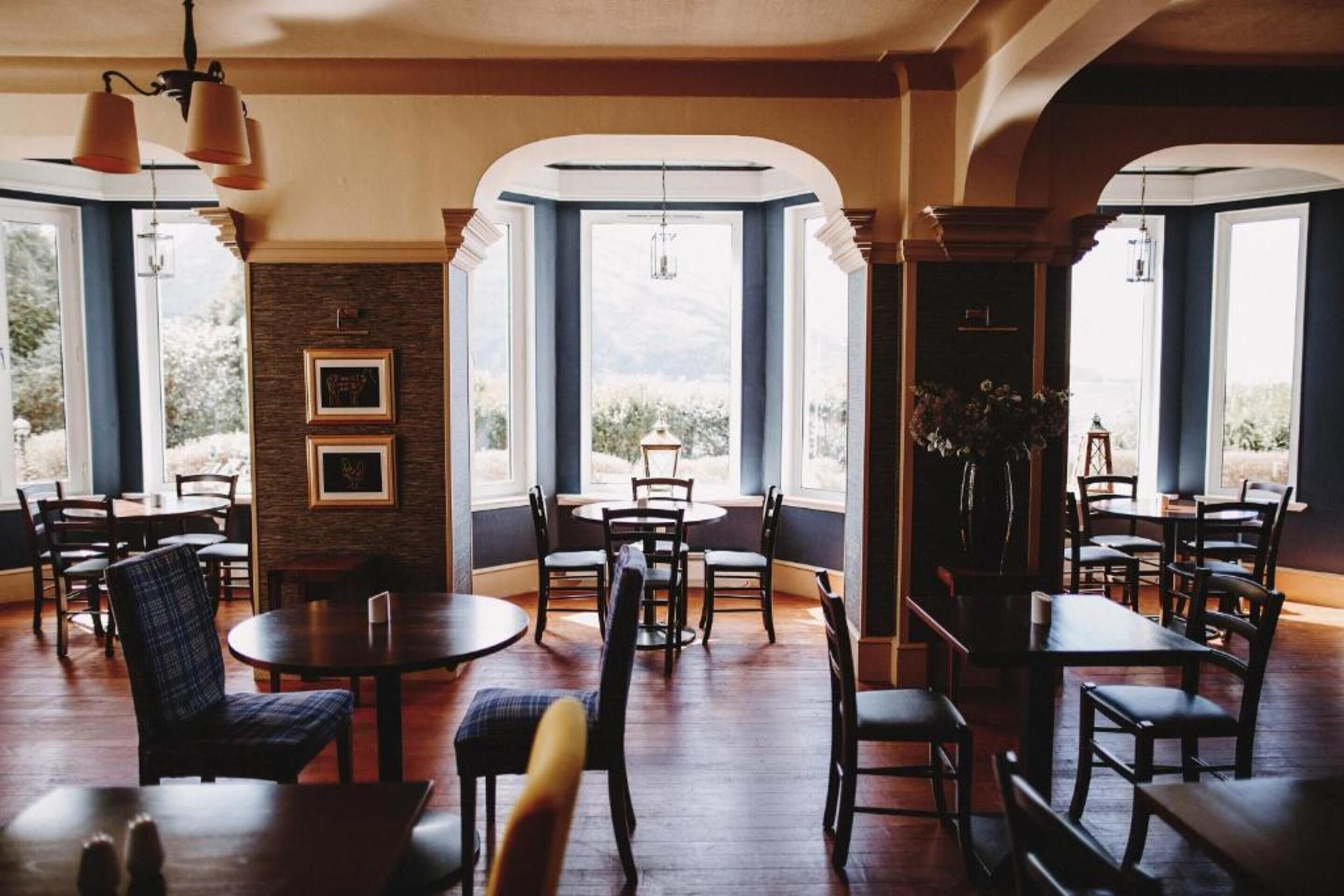 Un comedor con mesas y sillas redondas, ventanas amplias con vistas, y cuadros en la pared.