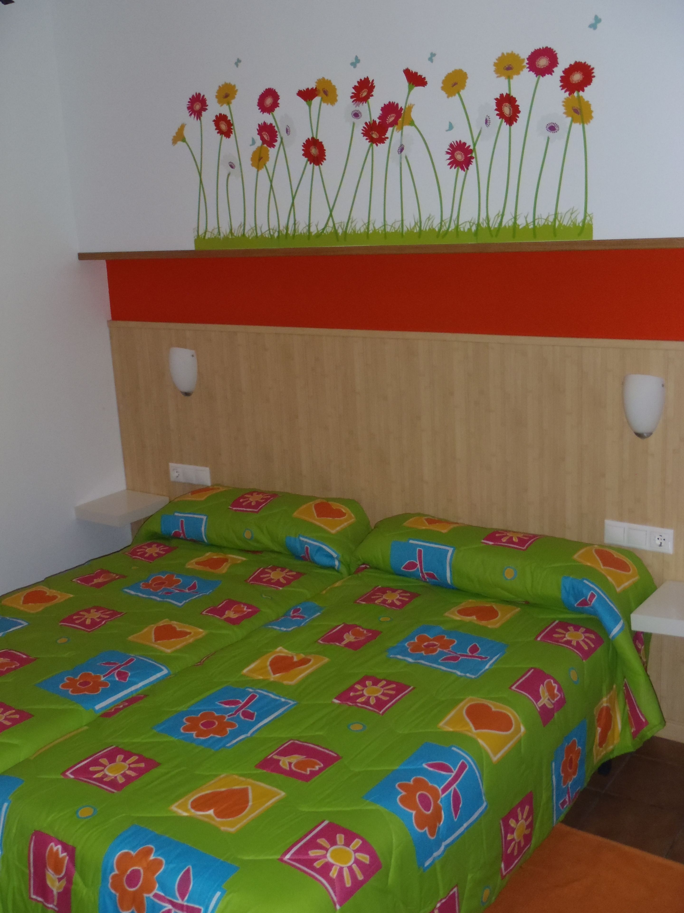 Una habitación con una cama, ropa de cama verde estampada, una pared con un mural floral y un estante rojo.