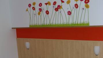 Una habitación con una cama, ropa de cama verde estampada, una pared con un mural floral y un estante rojo.