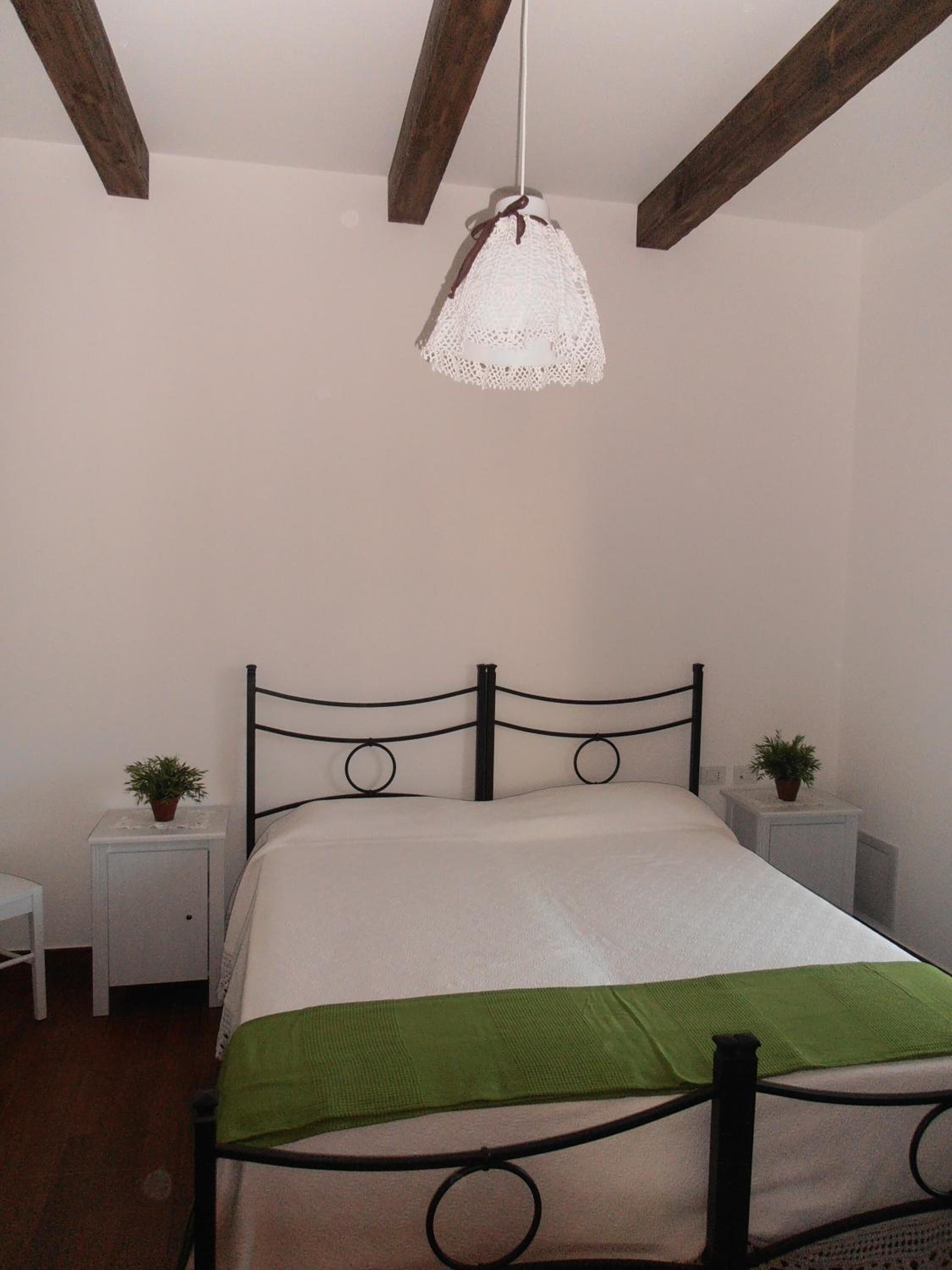 A bedroom with a bed, a white wall, a wooden ceiling beam, a hanging light fixture, and two bedside tables with plants.