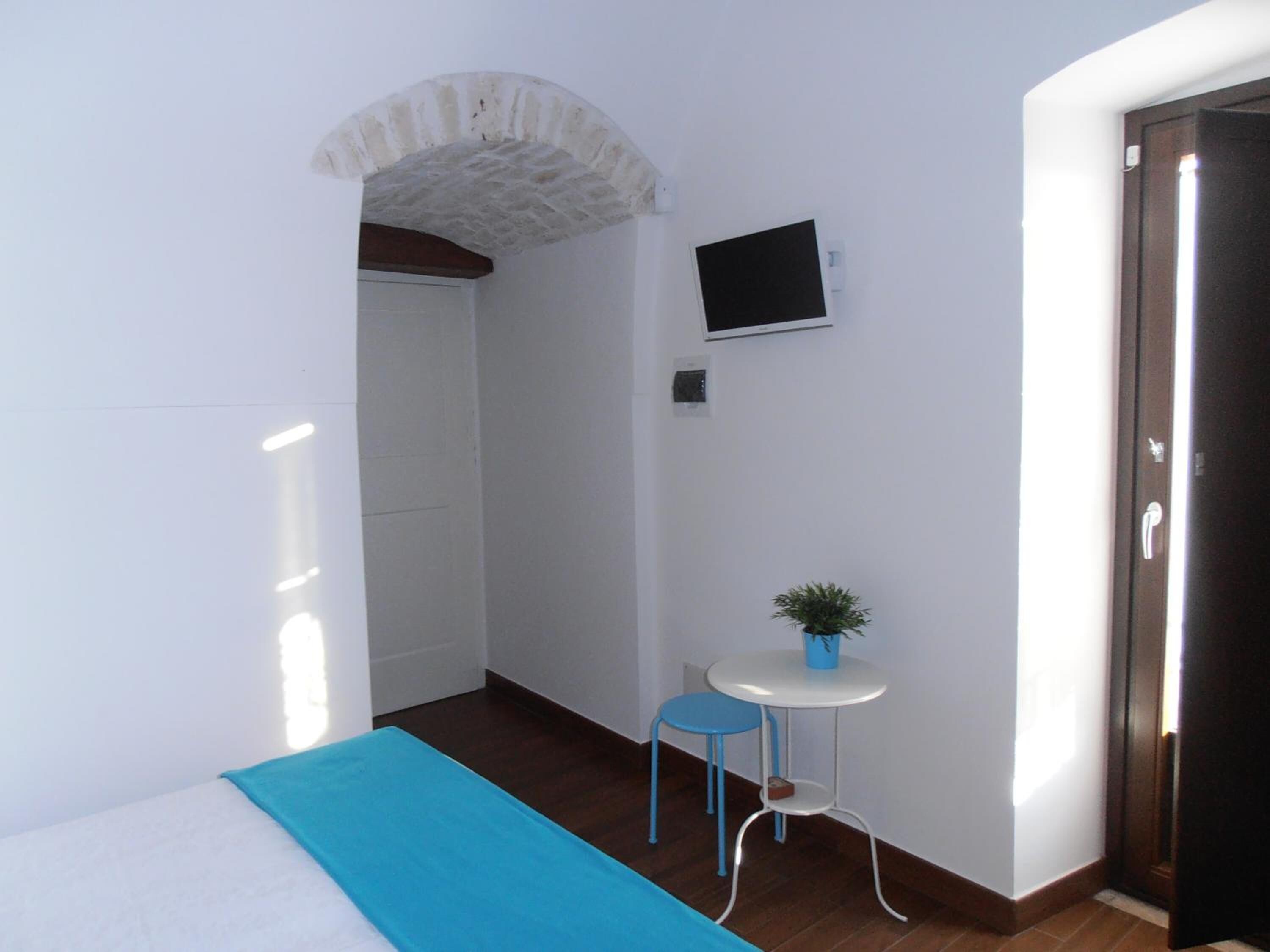 A small, bright room with a bed, a TV mounted on the wall, a small table with a plant, and a door.