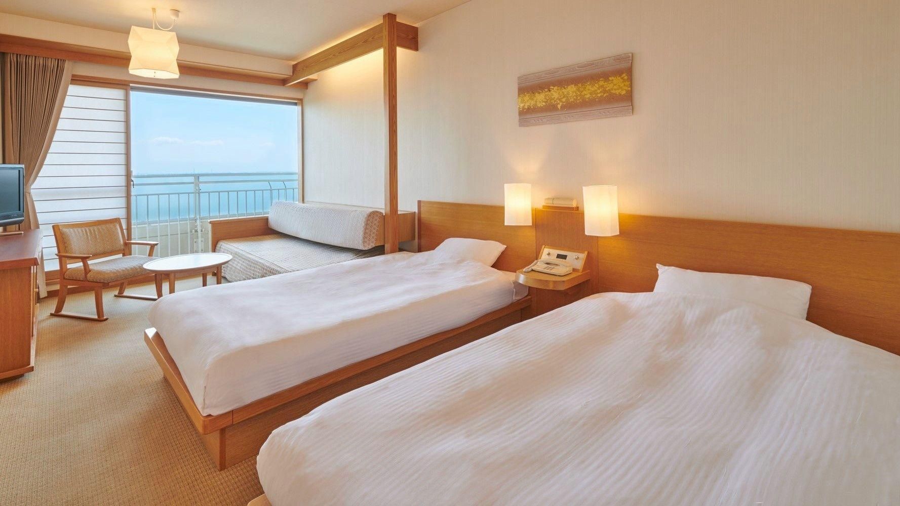 A hotel room with two beds, a desk, a chair, a small table, and a view of the sea.