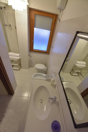 Un baño con bañera, lavamanos e inodoro. Hay una ventana sobre el inodoro y un perchero con toallas.