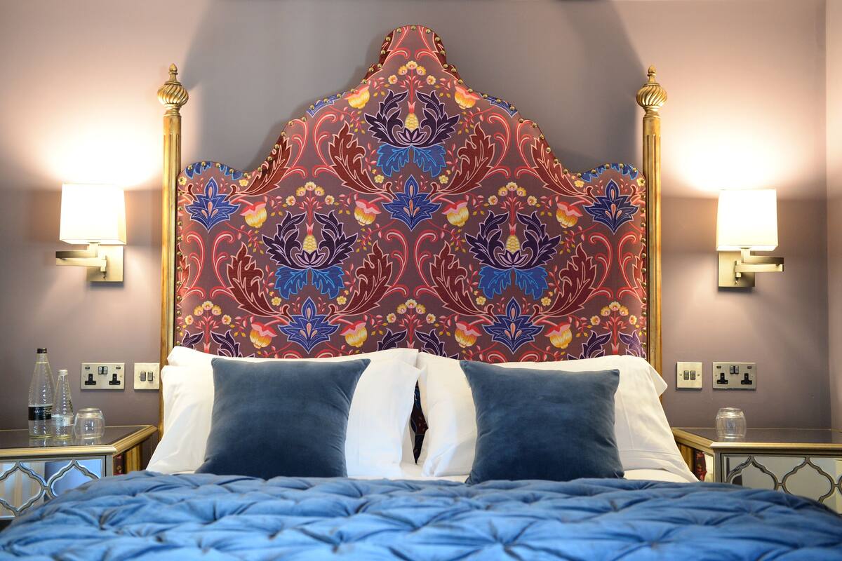 A bed with a blue quilt, a decorative headboard with floral patterns, two bedside lamps, and a nightstand with a bottle and glasses.