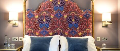 A bed with a blue quilt, a decorative headboard with floral patterns, two bedside lamps, and a nightstand with a bottle and glasses.