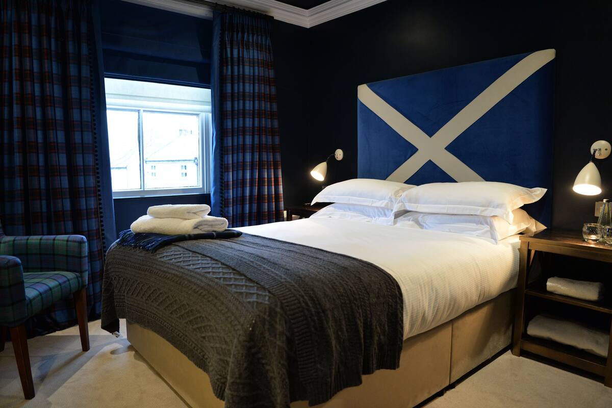 A hotel room with a large bed, a plaid chair, a window with curtains, and a wall with a Scottish flag design.