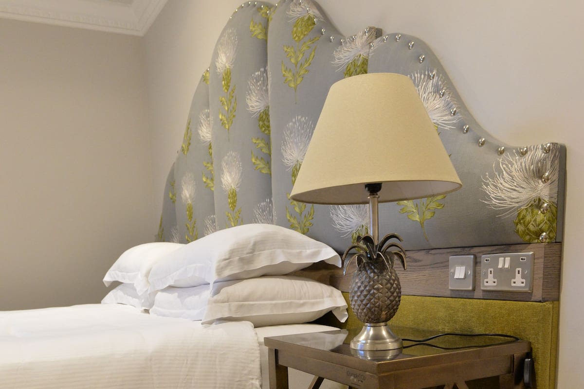 A neatly made bed with white linens, a lamp on a bedside table, and a plaid chair in the foreground.
