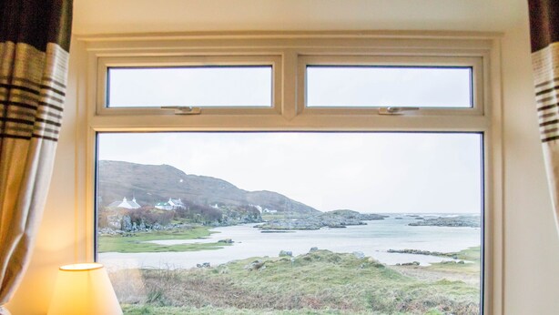 A room with a large window overlooking a coastal landscape, a lamp on a bedside table, and a chair. - Steading Holidays - Bungalow (Acharacle)