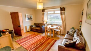 A living room with a brown leather sofa, a wooden dining table with chairs, a red striped rug, and a window with curtains. - Steading Holidays - Bungalow (Acharacle)