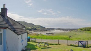 A coastal house with a grassy yard, a picnic table, and a view of the beach and hills. - Steading Holidays - Bungalow (Acharacle)