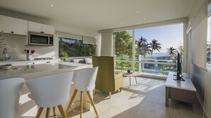 A modern kitchen with a central island, white chairs, a sofa, and a view of palm trees through large windows.