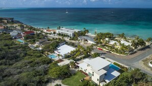 An aerial view of a coastal residential area with houses, a swimming pool, and a road leading to the beach.