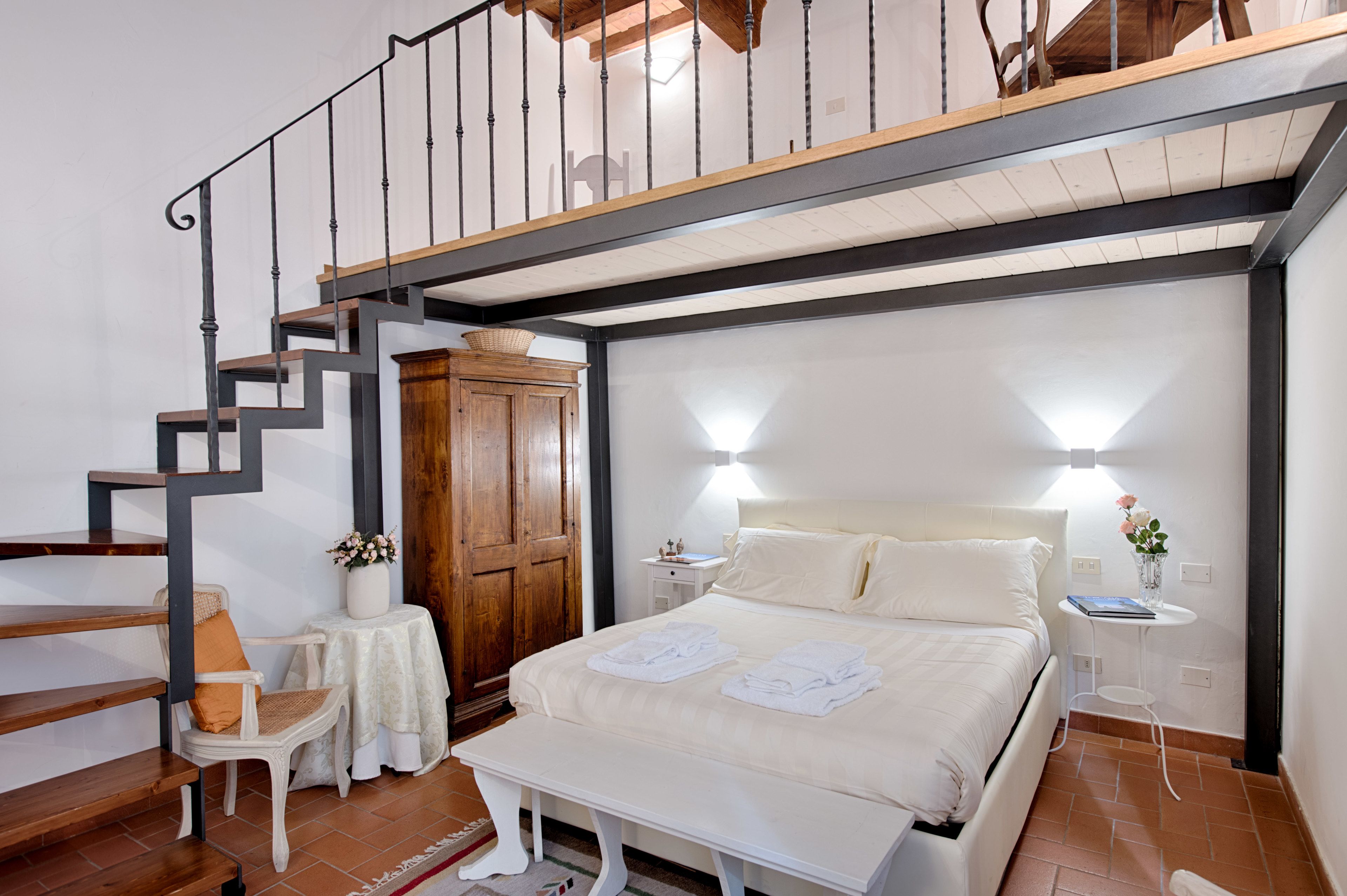 Un dormitorio con cama, mesitas de noche, armario de madera, una silla, una mesita con un florero y una escalera.