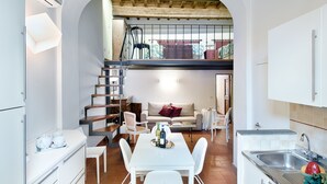 Une cuisine compacte avec des meubles blancs, un coin repas avec des chaises blanches, un escalier menant à une mezzanine et une vue sur l’extérieur à travers une arche.
