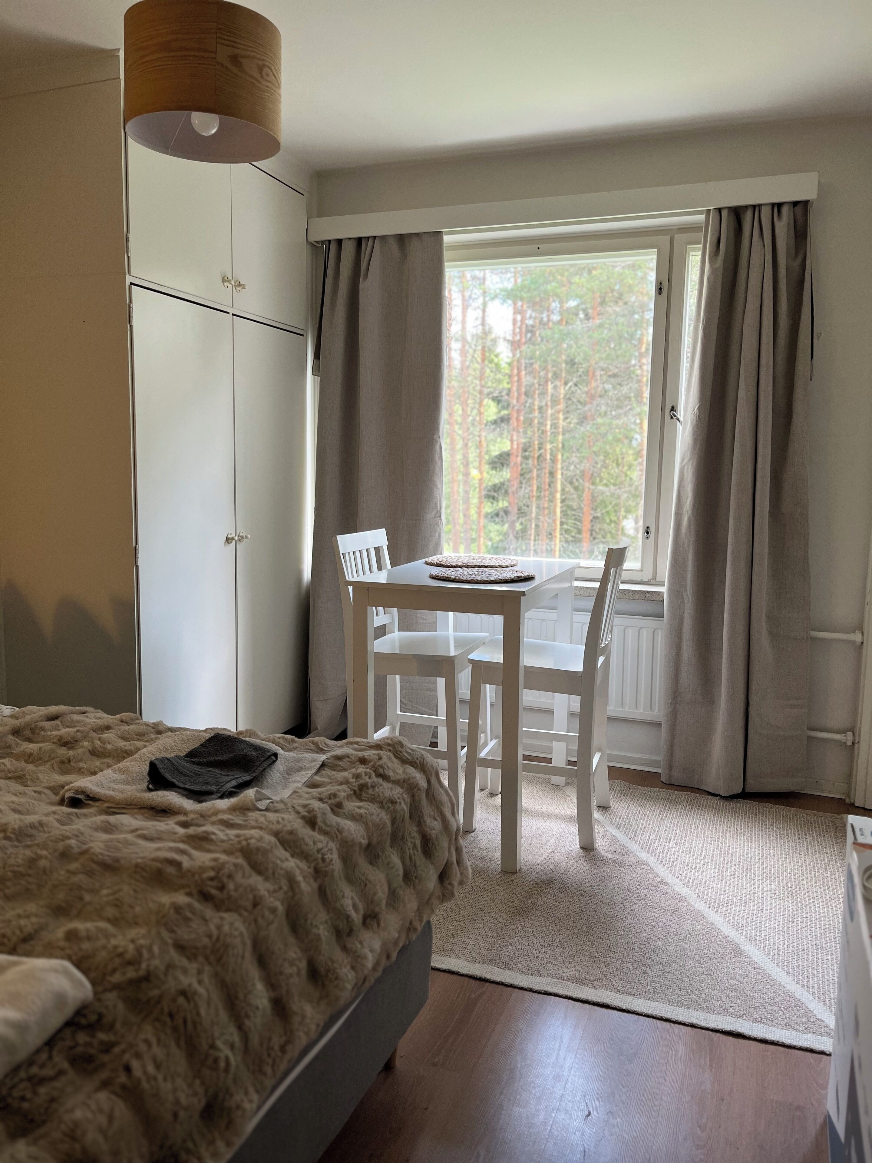 Un dormitorio con cama, armario, ventana con cortinas, mesa con sillas y vistas a árboles.