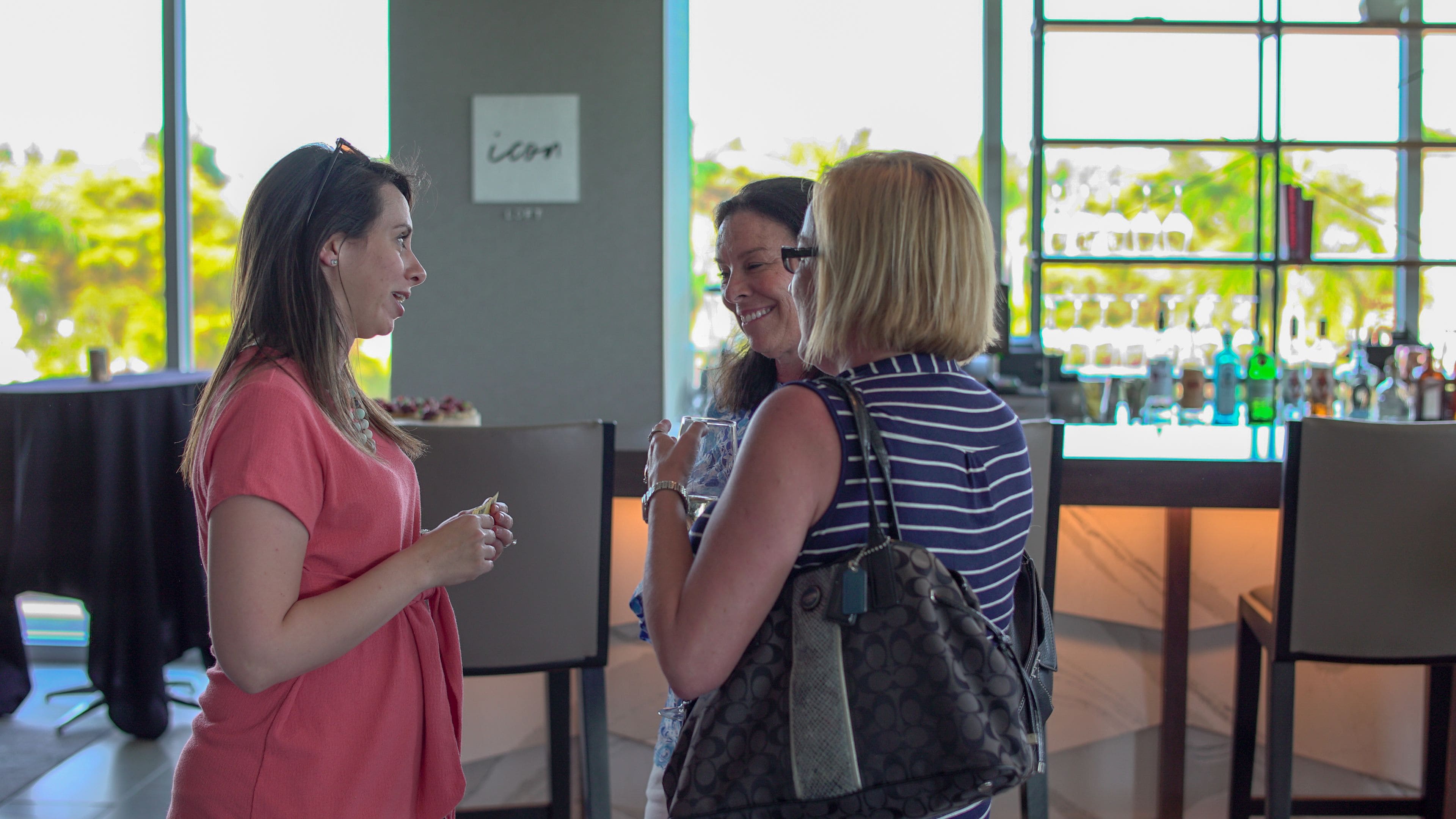 Three people conversing in a well-lit indoor space with large windows, a bar area, and a visible sign on the wall.