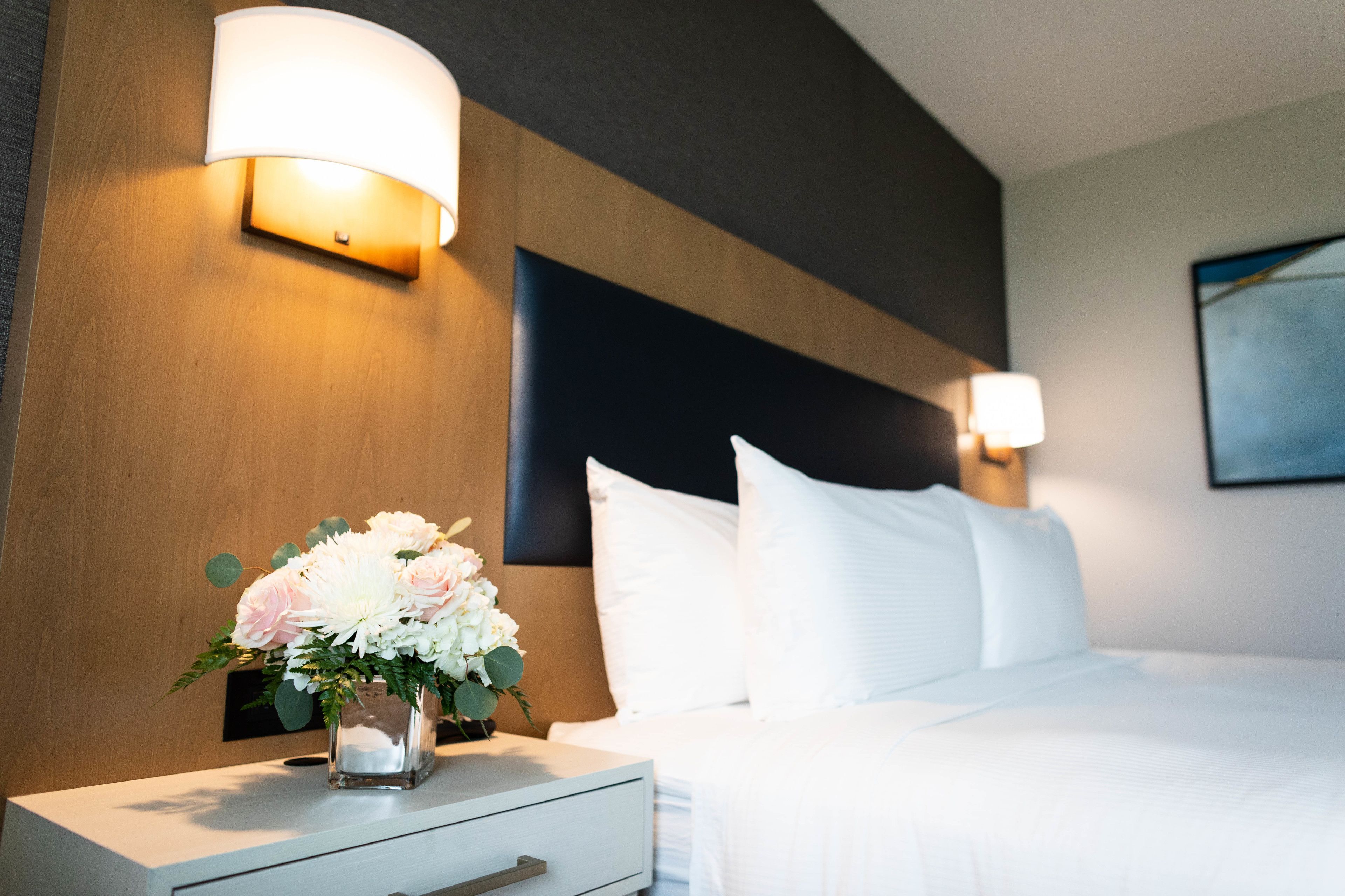 A hotel room with a neatly made bed, a bedside table with a vase of flowers, and wall-mounted lamps.