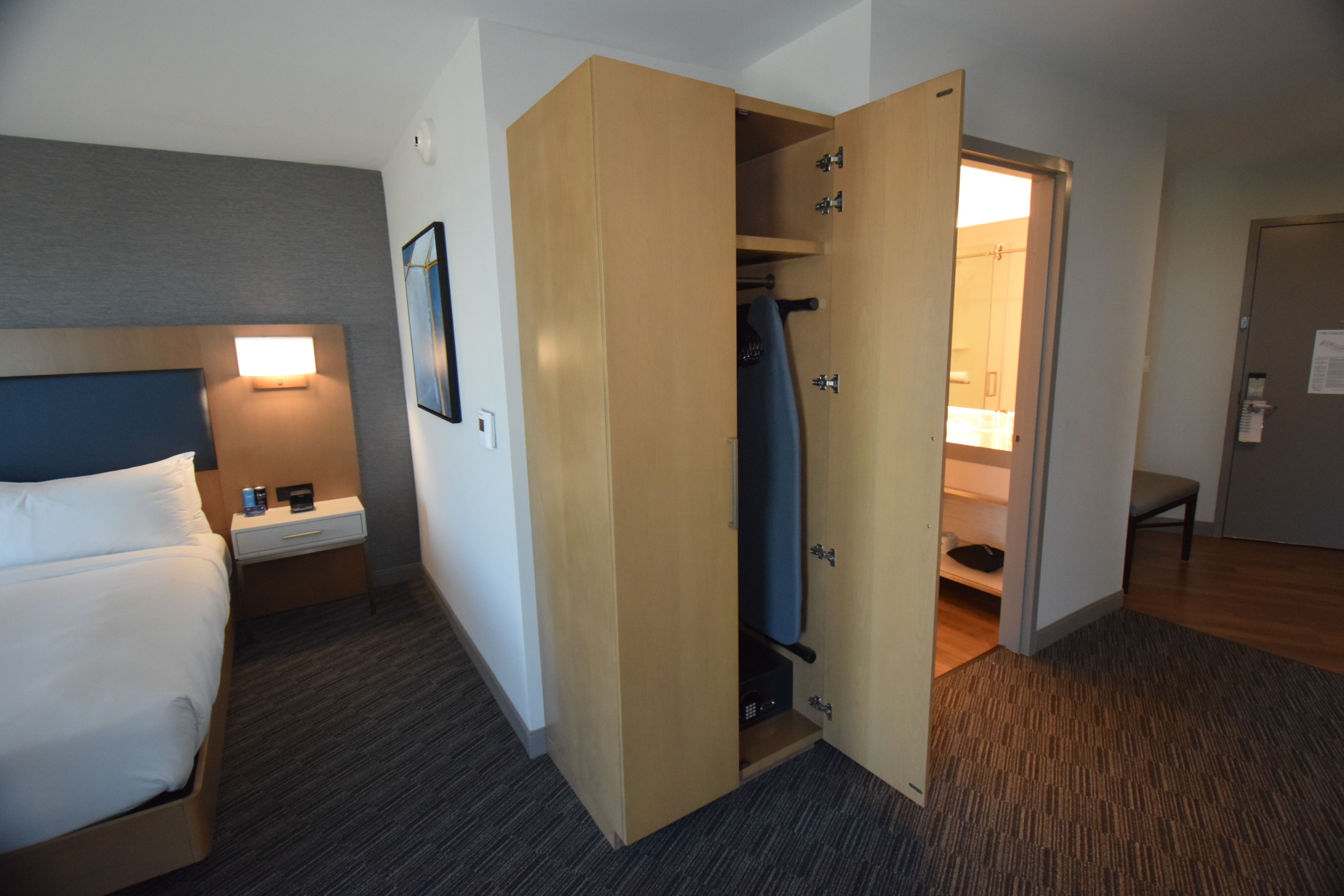 Habitación de hotel con cama, mesita de noche, armario y baño visible a través de una puerta abierta.