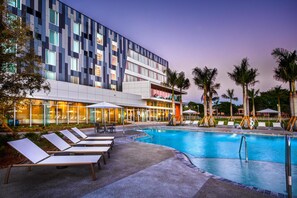 A modern hotel with a swimming pool, sun loungers, and palm trees.