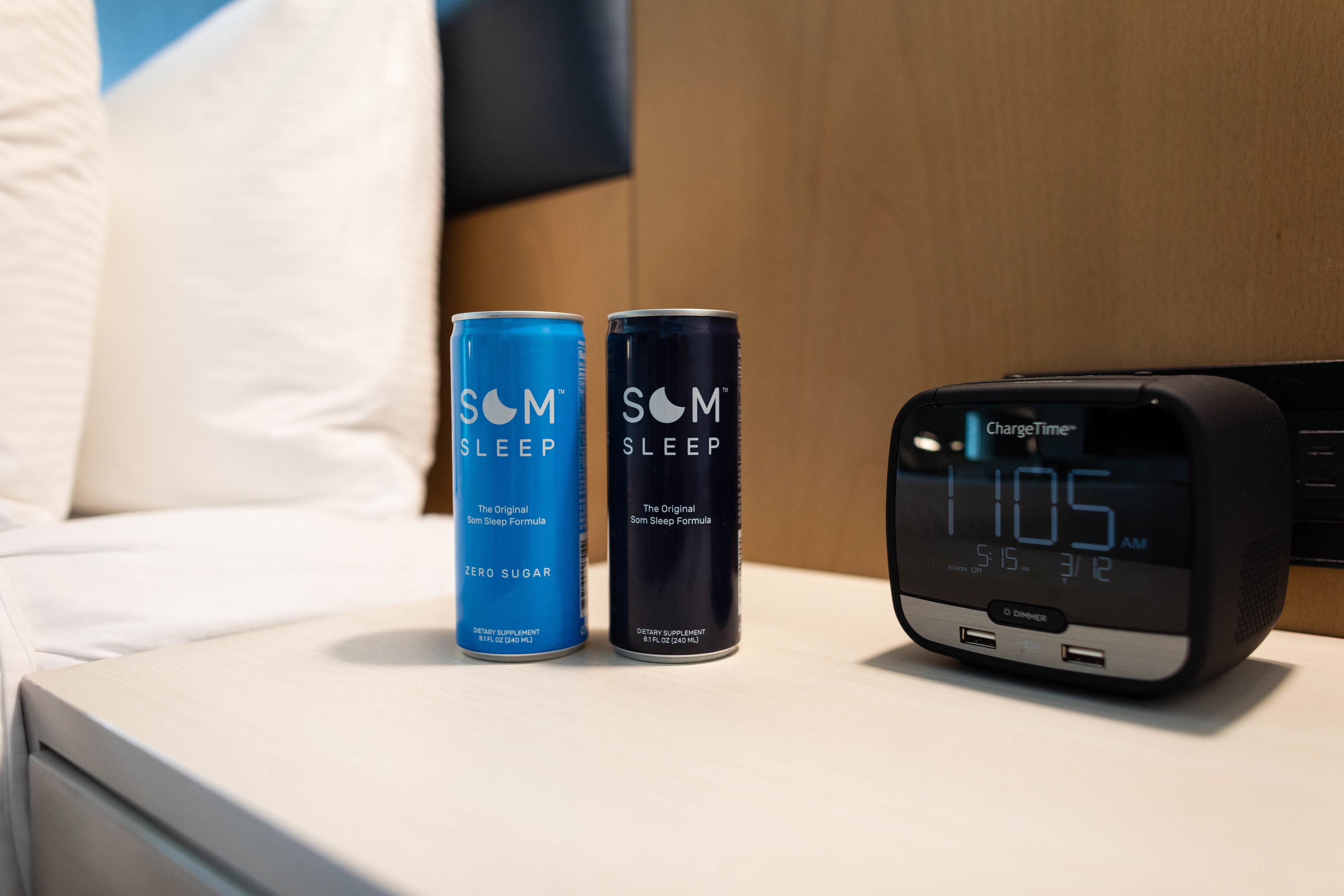 Una mesita de noche con dos latas de SOM Sleep Formula y un reloj digital ChargeTime que marca las 11:05 AM.