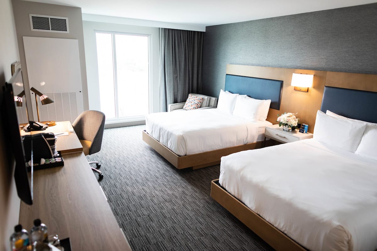 A hotel room with two beds, a desk with a lamp, a chair, a dresser, and a large window with curtains.