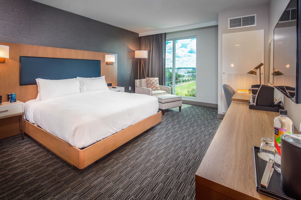 A hotel room with a large bed, a desk with a chair, a lamp, and a view of the outdoors.