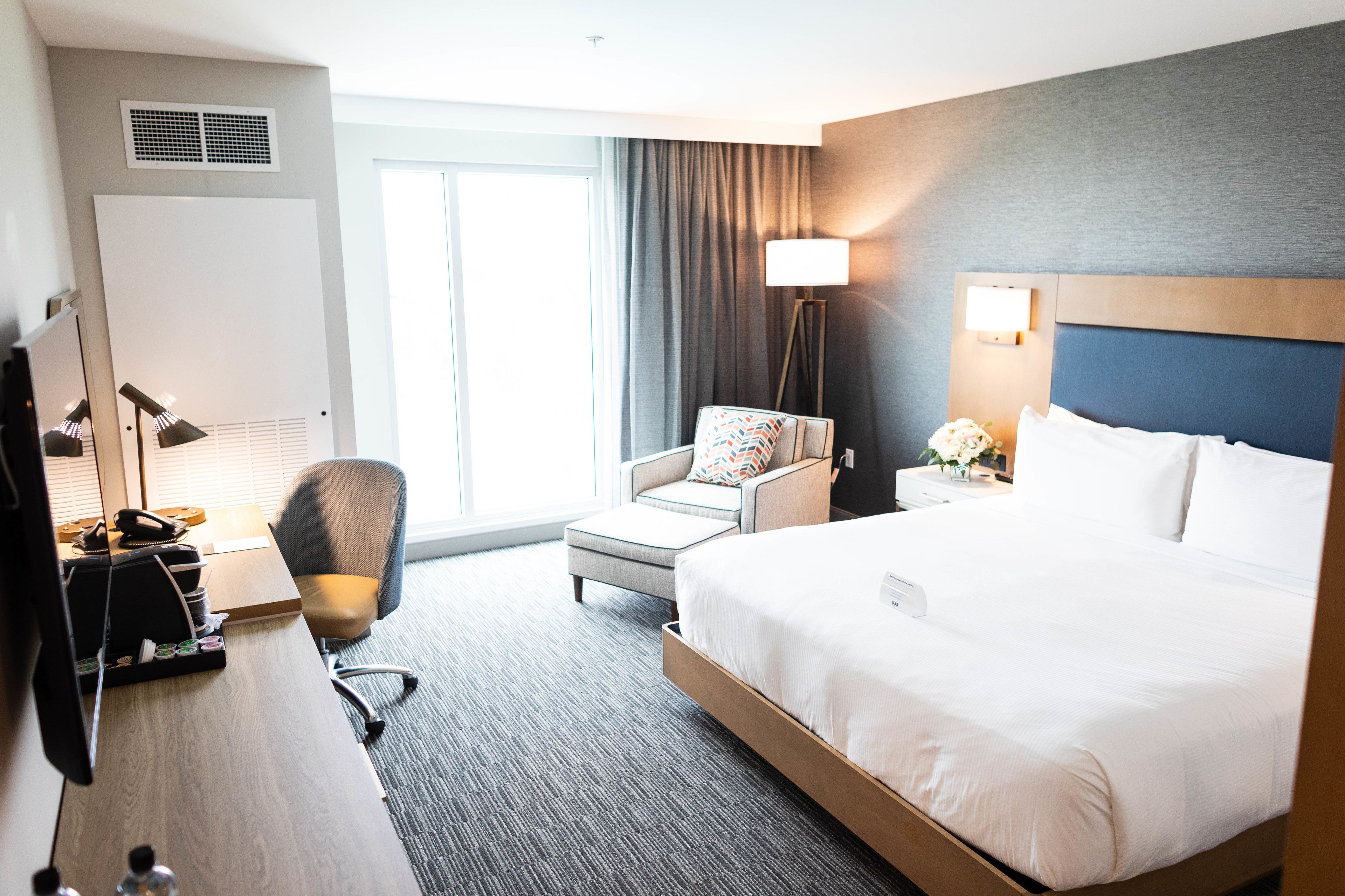 A hotel room with a large bed, a desk with a computer, a chair, a lamp, and a window with curtains.