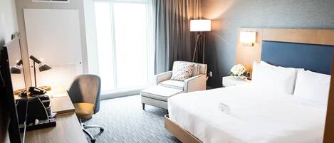 A hotel room with a large bed, a desk with a computer, a chair, a lamp, and a window with curtains.
