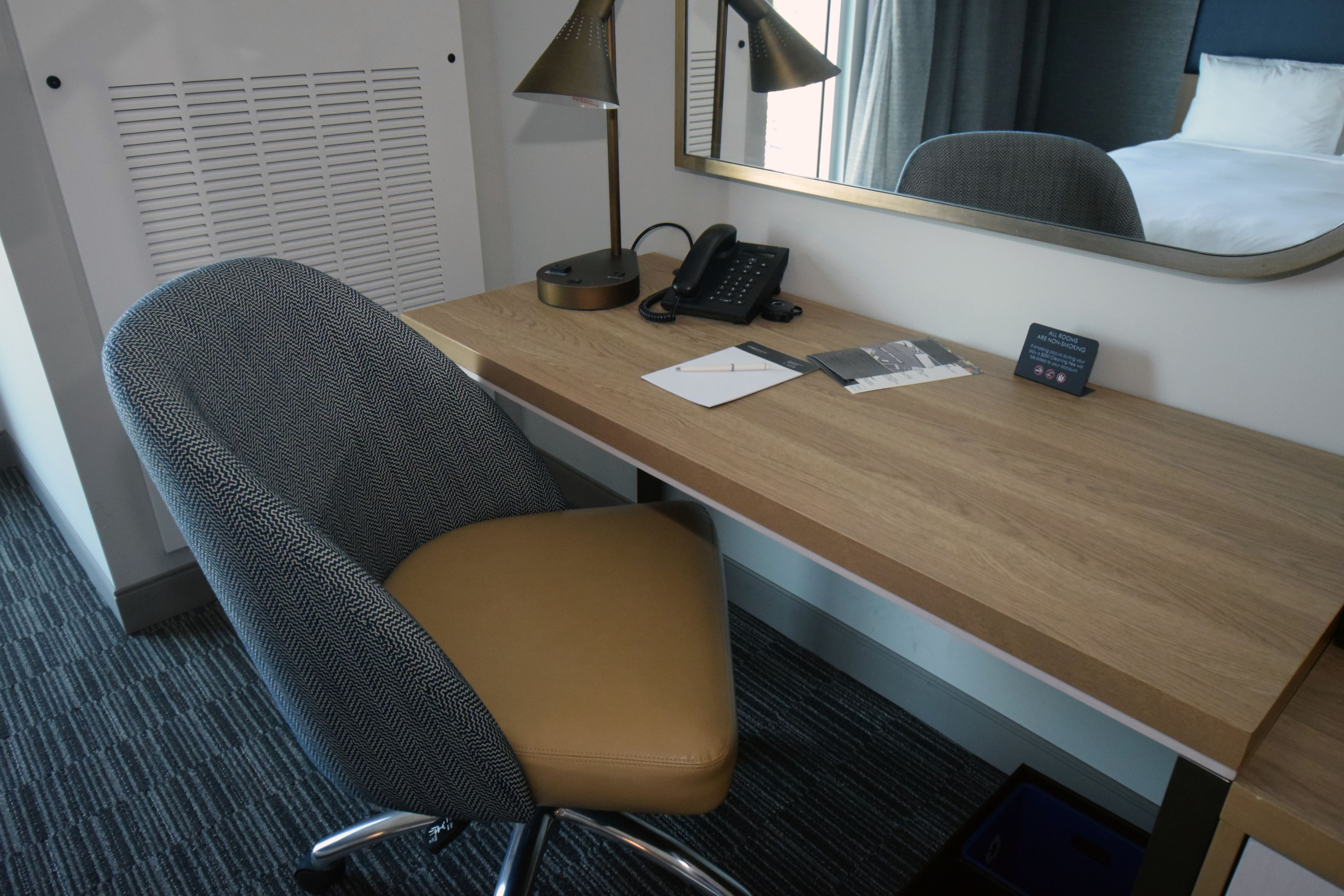 Une chambre d’hôtel avec un bureau en bois, un téléphone, une lampe, une chaise et un lit doté d’une tête de lit.