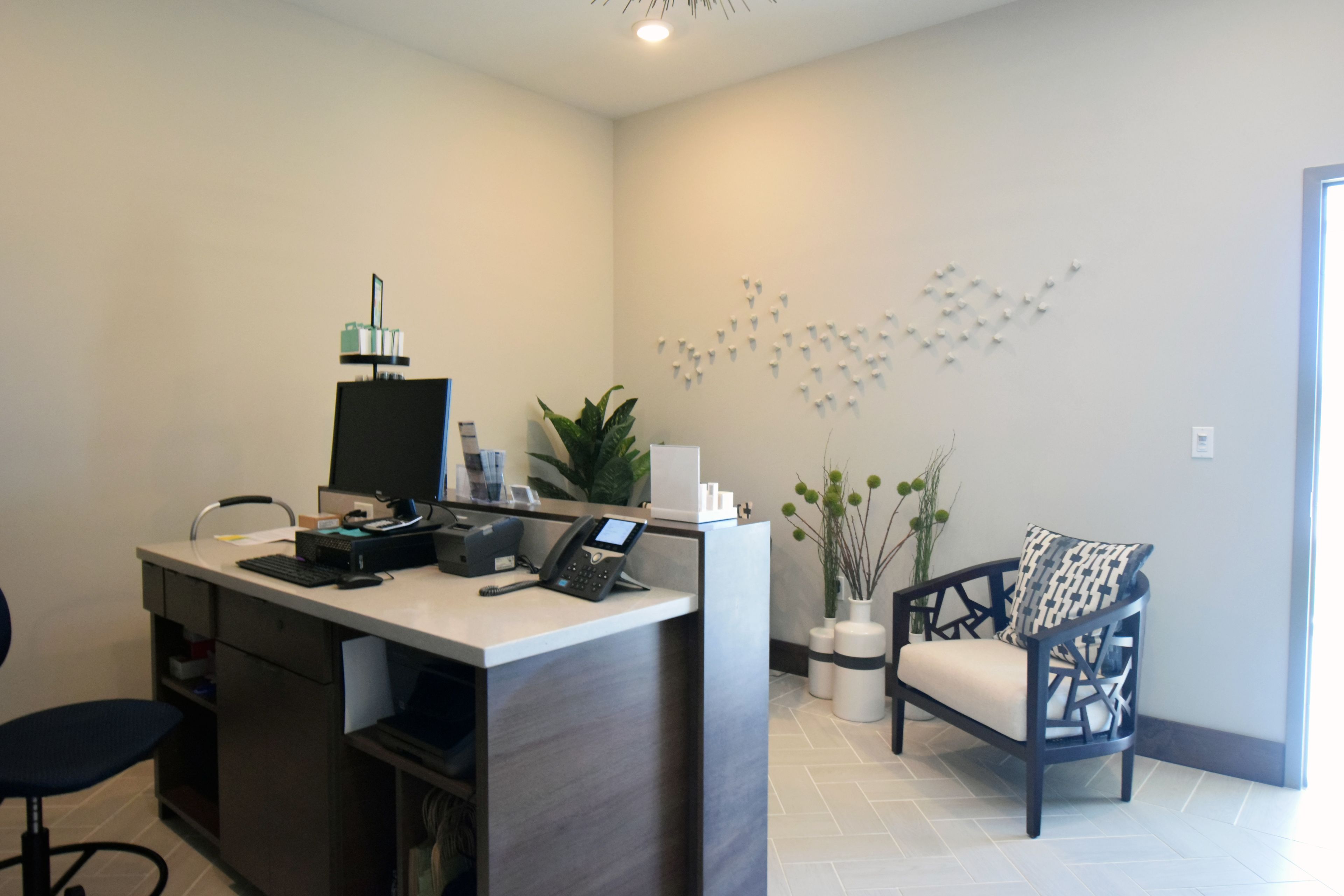 A reception desk with a computer, phone, and printer, a chair, a plant, and a wall-mounted art piece.