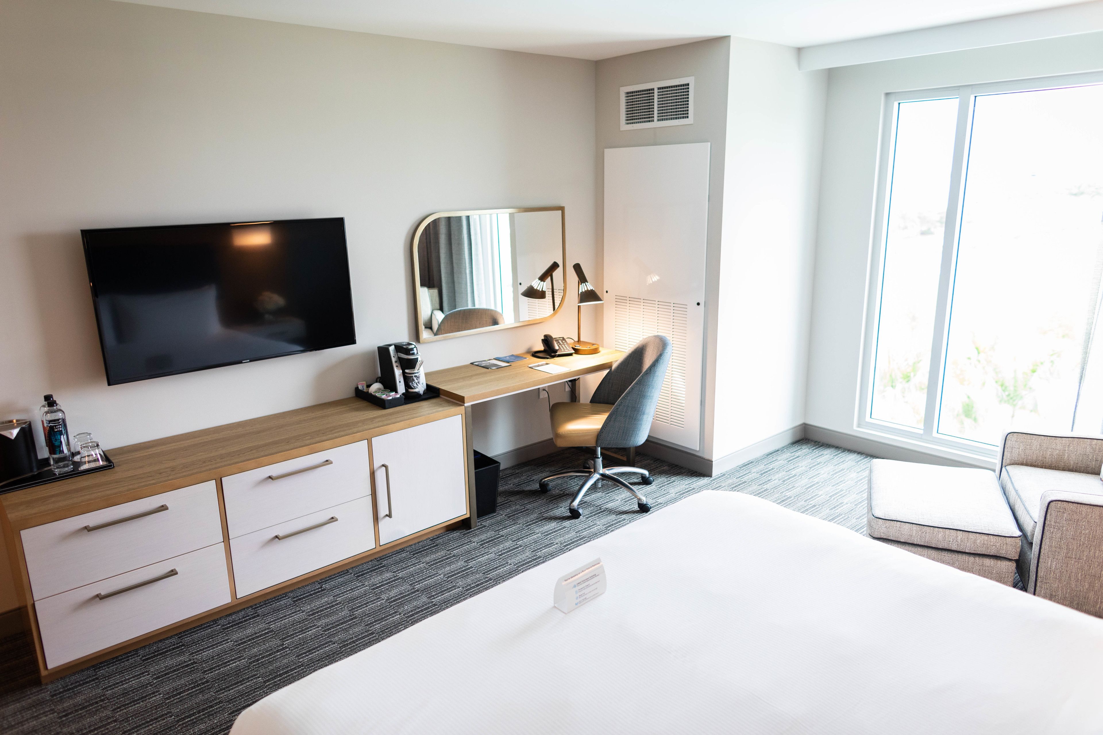 A hotel room with a bed, a desk with a chair, a TV, a mirror, and a wardrobe.