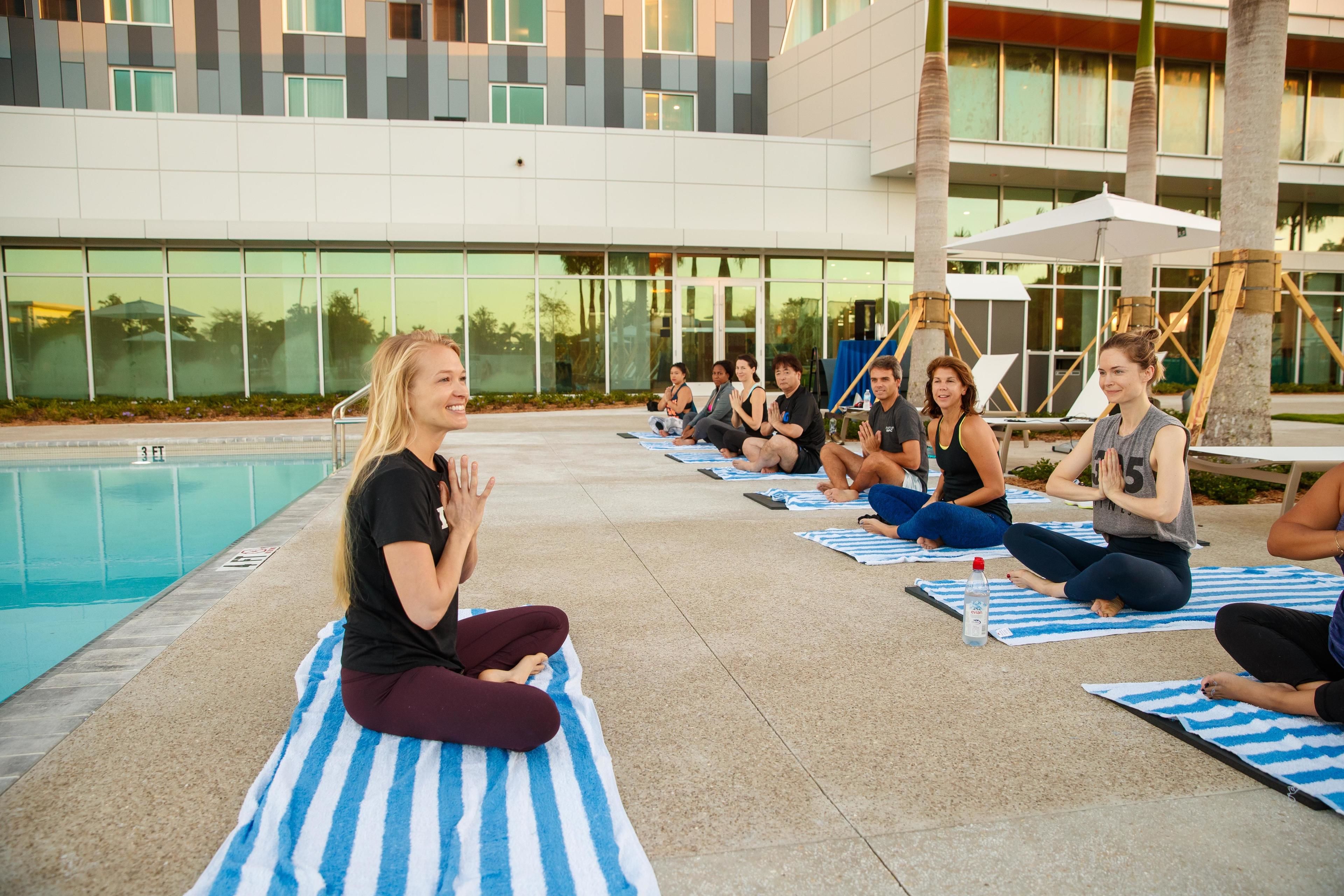 Een groep mensen zit op yogamatten in een gedeeltelijk openluchtruimte, met een zwembad op de achtergrond.