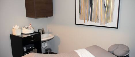 A hotel room with a bed, a bedside table with a lamp, a sink, and a framed abstract painting on the wall.