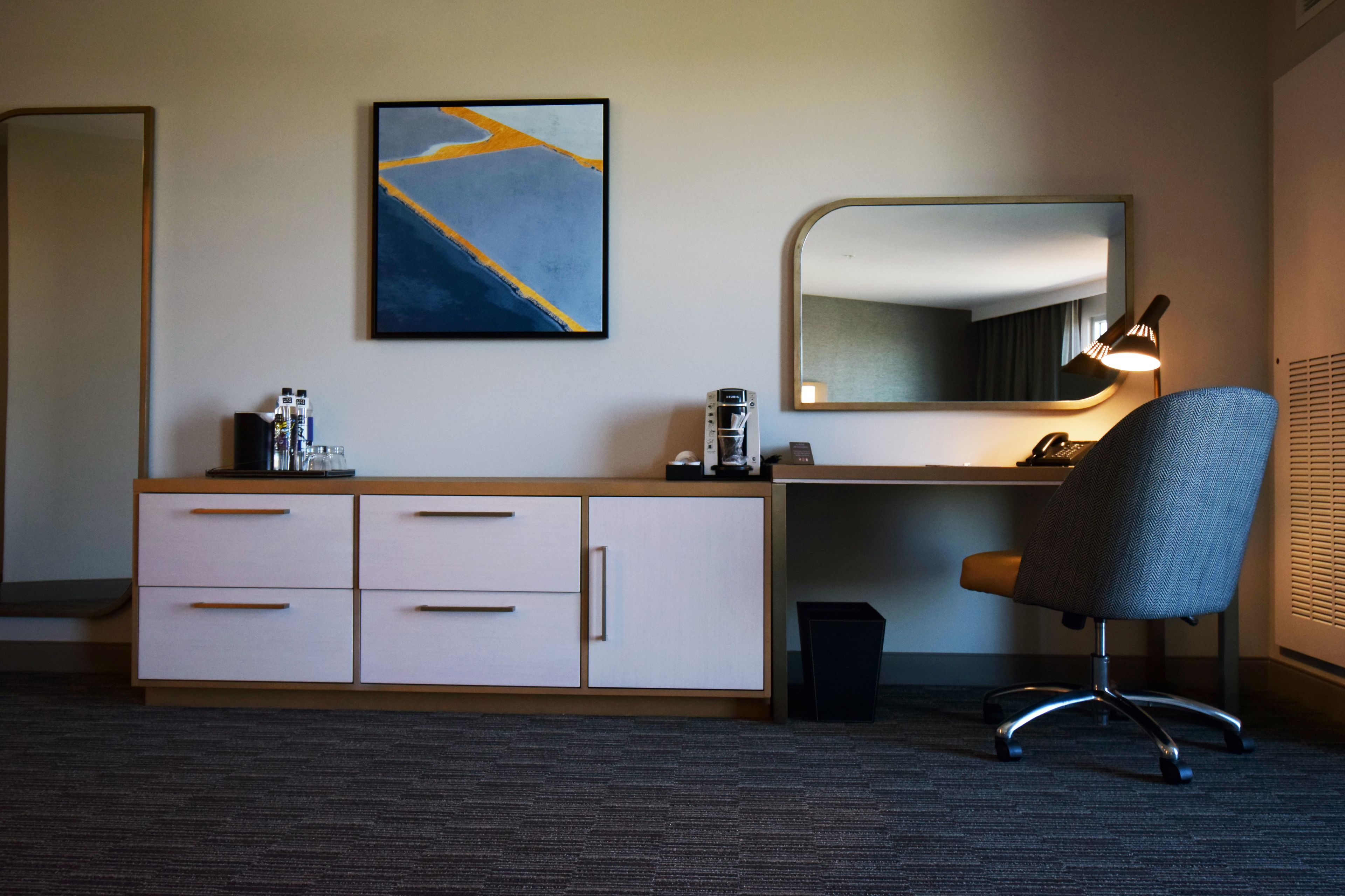 A hotel room with a desk, a chair, a large mirror, a small trash bin, and a wall-mounted artwork.