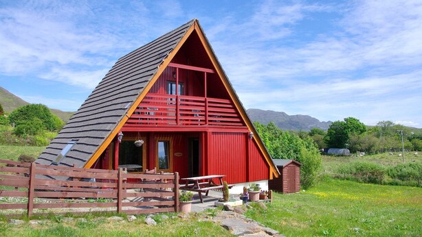 A red A-frame cabin with a wooden fence, a picnic table, and a small shed in a grassy field with mountains in the background. - Steading Holidays - Comraich (Acharacle)