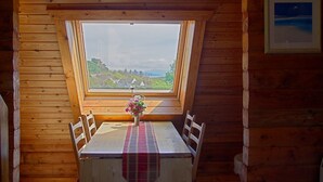 A wooden cabin with a dining area featuring a table, chairs, and a view through a large window. - Steading Holidays - Comraich (Acharacle)