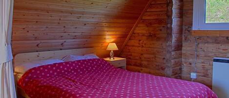 A cozy bedroom with a wooden sloped ceiling, a single bed with a red patterned bedspread, a bedside table with a lamp, and a window with a view of greenery.