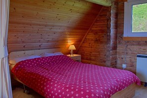 A cozy bedroom with a wooden sloped ceiling, a single bed with a red patterned bedspread, a bedside table with a lamp, and a window with a view of greenery. - Steading Holidays - Comraich (Acharacle)