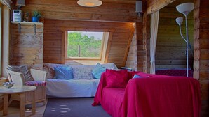 A cozy cabin interior with a bed, a sofa, a dining table, and a window offering a view of greenery. - Steading Holidays - Comraich (Acharacle)