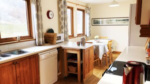 A compact kitchen with wooden cabinets, a dishwasher, a microwave, and a dining area with a table and chairs. - Steading Holidays - Eilean Feoir (Acharacle)