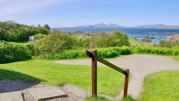 A gravel path leading to a wooden gate, surrounded by green grass and bushes, with a view of the sea and distant mountains. - Steading Holidays - Eilean Feoir (Acharacle)