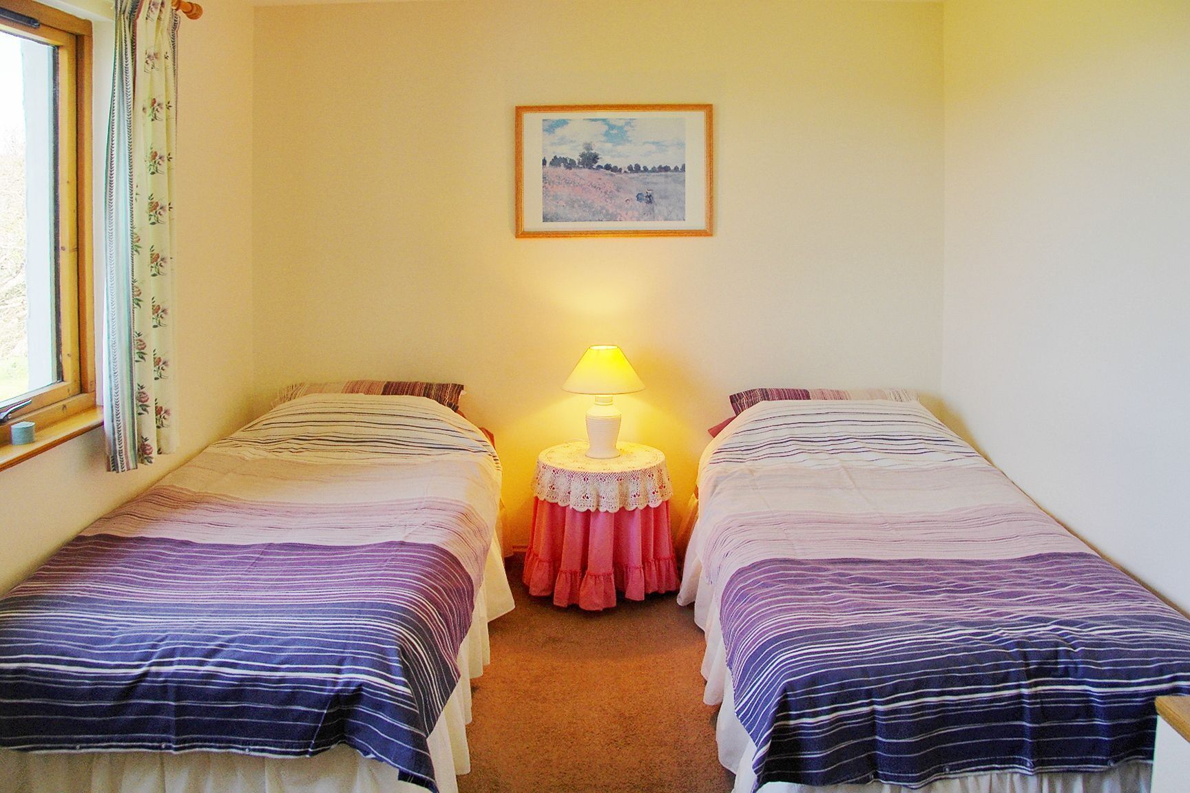 A small room with two single beds, a bedside table with a lamp, a framed picture on the wall, and a window with curtains.