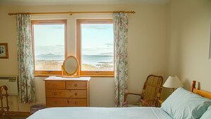 A bedroom with a bed, dresser, chair, and a window with a view of the beach. - Steading Holidays - Eilean Feoir (Acharacle)