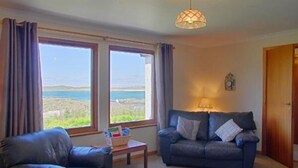 A living room with a sofa, a chair, a table with books, and a lamp. There are large windows with curtains and a view of the sea. - Steading Holidays - Eilean Feoir (Acharacle)